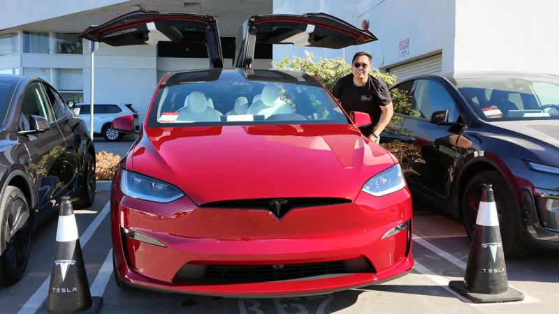 A man stands beside a red Tesla Model X with its falcon wing doors open in a parking lot.