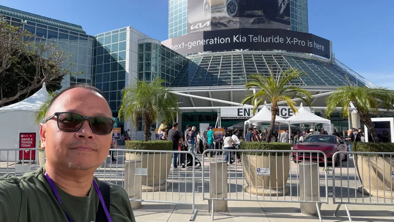 Man in sunglasses takes selfie at outdoor auto show entrance with convention center, Kia banner.