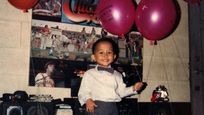 Happy young boy in a bowtie holds balloons in front of a band poster and speakers.