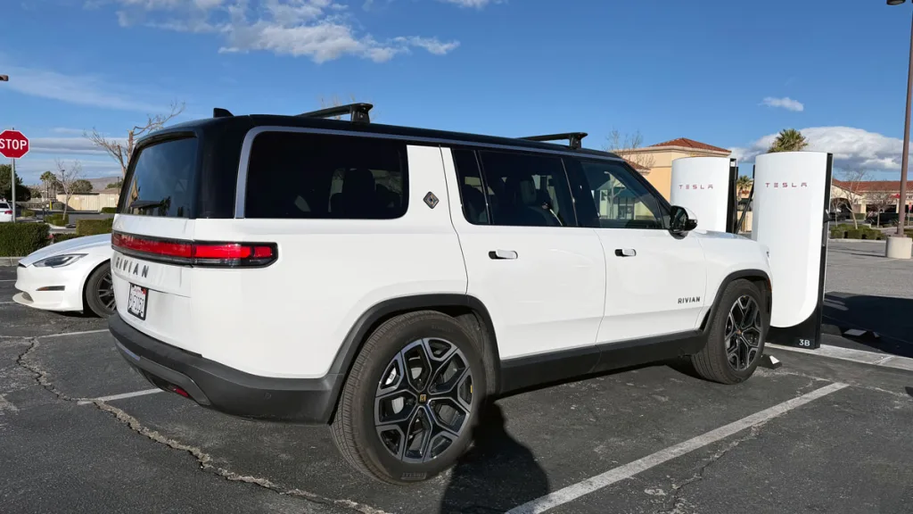 White Rivian R1S SUV parked near Tesla Superchargers with a white Tesla car behind it.