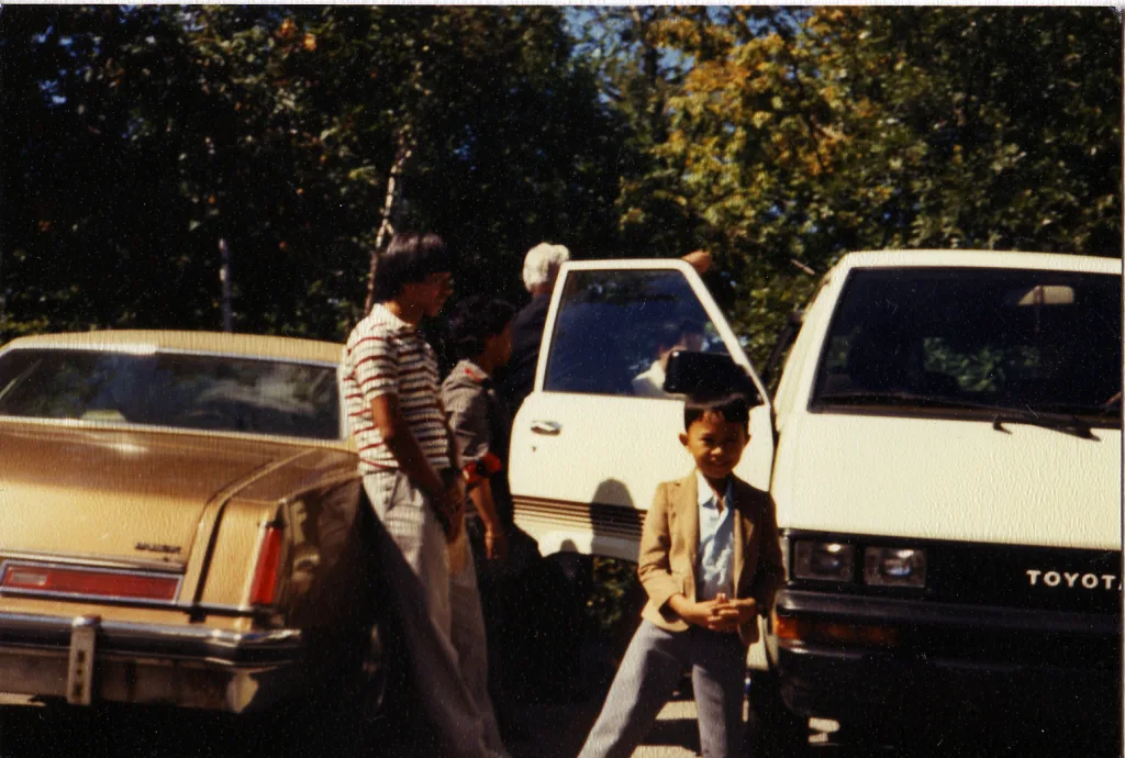Smiling boy in blazer with two men and vintage cars, a gold sedan and a white Toyota van.